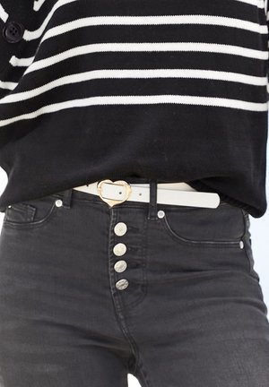 Person trägt einen schwarz-weiß gestreiften Pullover, gesteckt in eine schwarze Jeans mit silbernen Knöpfen und einem weißen Gürtel mit goldener herzf örmiger Schnalle.