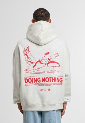 Sudadera gris con un gran gráfico rojo en la parte trasera que representa figuras tumbadas y un texto que dice "NO HACIENDO NADA". Cuenta con capucha ajustable.