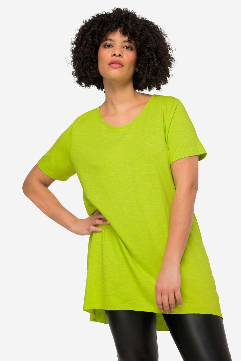 Femme aux cheveux bouclés portant un haut vert vif oversized à manches courtes et des leggings noirs, posant avec une main sur la hanche devant un fond uni.
