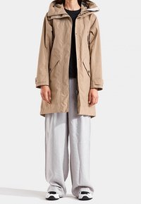 Beige vedenpitävä parkatakki hupulla, etuvetoketjulla, kahdella sivutaskulla ja väljä istuvuus, yhdistettynä löysiin, vaaleanharmaisiin housuihin ja tennareihin.