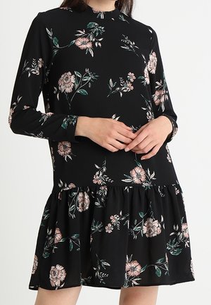 Černé šaty s dlouhými rukávy s růžovým květinovým potiskem, vysokým límcem a nařasenou sukní, nošené osobou s dlaněmi sepnutými před sebou.