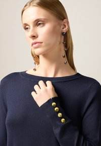 Maglione a maglia blu navy con scollo rotondo e dettagli con bottoni dorati sui polsini. Capelli lunghi e orecchini pendenti a motivo visibili.