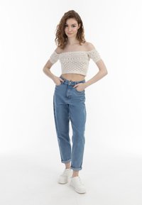 Světle béžový háčkovaný crop top s off-shoulder designem, zkombinovaný s vysokým pasem světle modrými džíny a bílými teniskami, stojící na bílém pozadí.