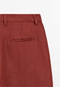 Gros plan de l'arrière d'un pantalon en lin couleur rouille avec passants de ceinture et une poche passepoilée unique sur fond blanc.