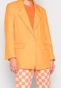 Oranger Blazer mit einem Knopfverschluss, Reverskragen und zwei vorderen Taschen, kombiniert mit rosa-weißen karierten Hosen.