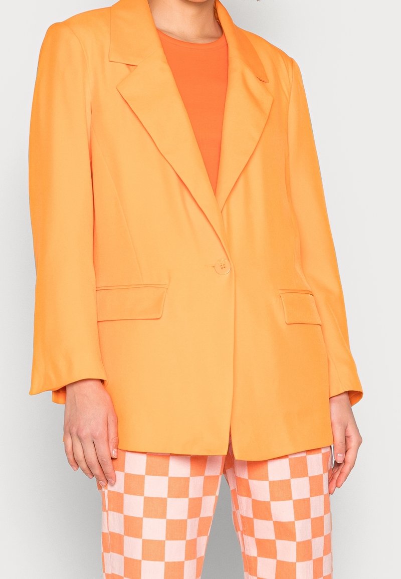 Oranger Blazer mit einem Knopfverschluss, Reverskragen und zwei vorderen Taschen, kombiniert mit rosa-weißen karierten Hosen.