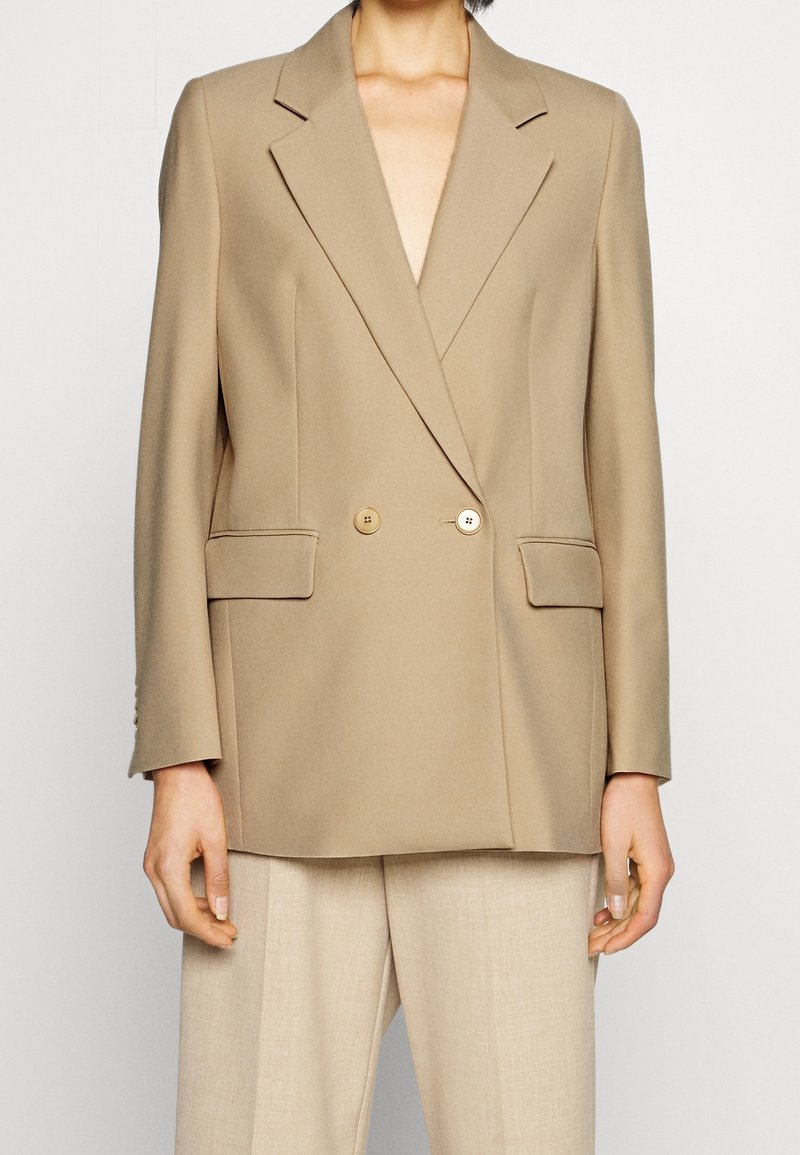 Blazer beige de doble botonadura con solapas de pico, dos bolsillos frontales y botones dorados, confeccionado en tejido texturizado. Visten unos pantalones beige.