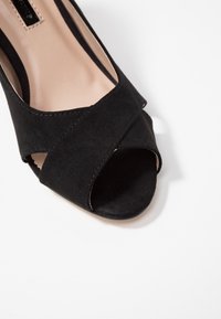 Černé zamšové otevřené podpatky s překříženými pásky. Hladký povrch, zakřivený tvar a neutrální vnitřní podšívka. Prošití podél okrajů.