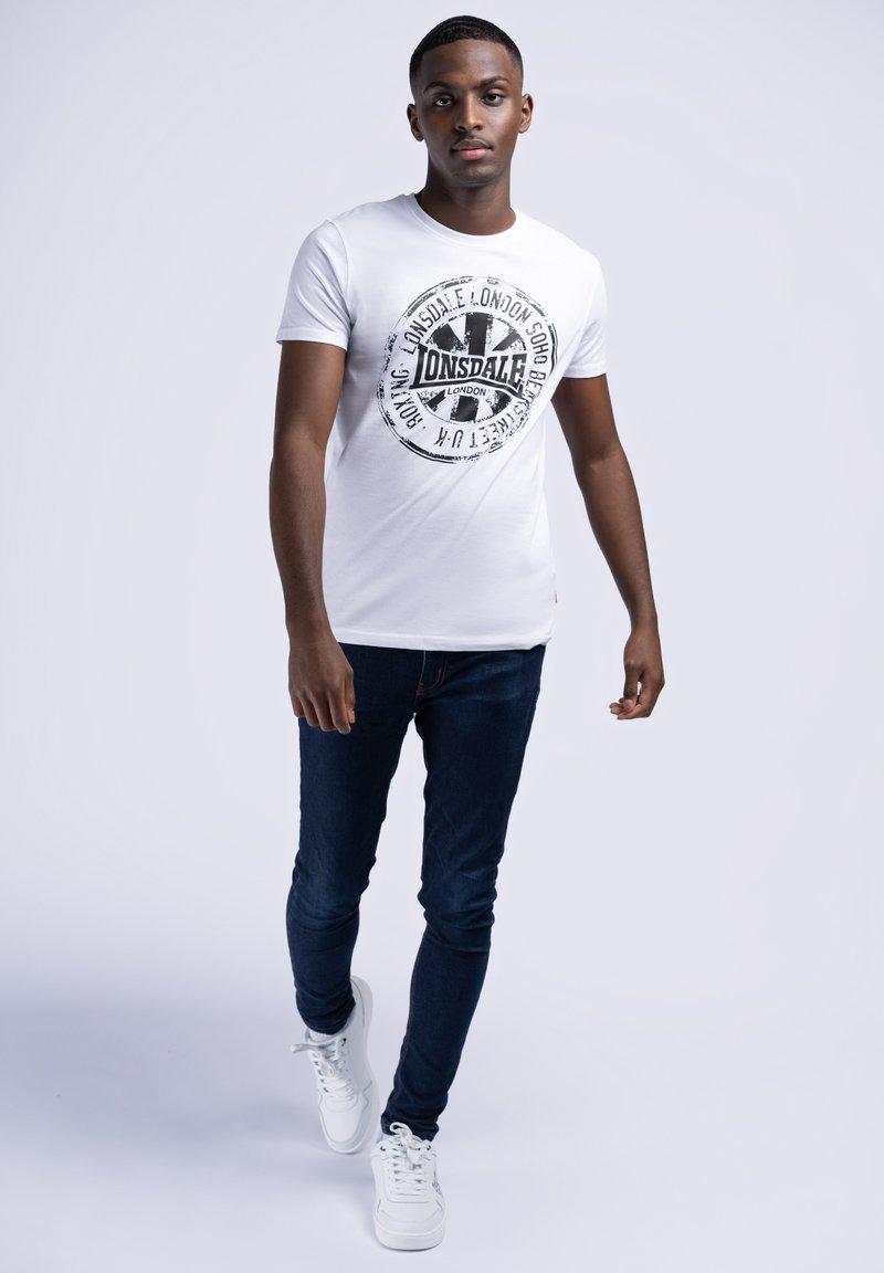 Tricou alb cu imprimeu grafic negru al logo-ului Lonsdale, asociat cu blugi skinny închiși la culoare și adidași albi.