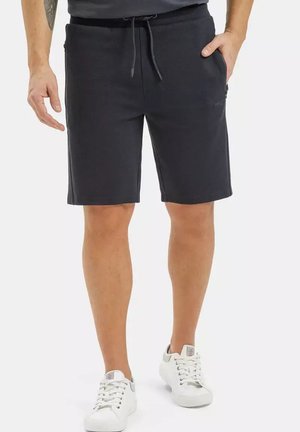 Mężczyzna noszący czarne szorty do kolan z wiązanym paskiem w talii i białe trampki, stojący z jedną ręką w kieszeni.