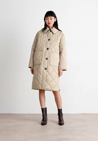 Manteau beige matelassé avec col montant, fermeture à boutons et grandes poches. Présente une coupe ample et un design mi-long. Texture unie.