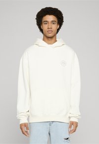 Sudadera con capucha color crema confeccionada en tela suave, con bolsillo delantero, capucha con cordón y un parche con logo redondo en el pecho. Se lleva con vaqueros azul claro.