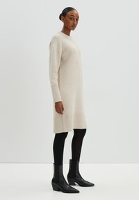 Robe pull beige en maille avec des poignets et un ourlet côtelé, associée à des bottines noires élégantes et des collants noirs, sur un fond neutre.