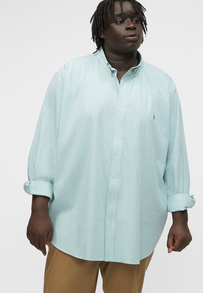 Osoba tamne puti i ispletene kose, odjevena u široku, svijetlozelenu i bijelu prugastu košulju s gumbima i bež hlače, stoji na jednostavnoj pozadini.