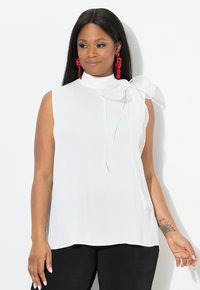 Weiße ärmellose Bluse mit hohem Kragen und großem transparentem Bogen auf der linken Schulter. Glatte Stoffqualität, kombiniert mit schwarzen Hosen.