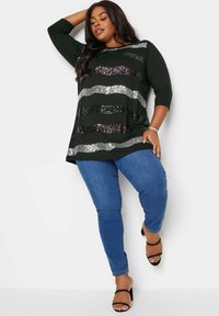 Schwarzes Oberteil mit kräftigen horizontalen Streifen aus Silber- und tiefvioletten Pailletten, kombiniert mit blauen engen Jeans und schwarzen High Heels.