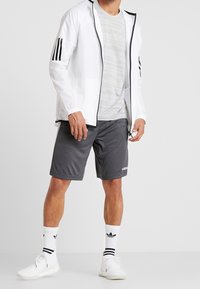 Chaqueta blanca ligera con detalles en negro y tres rayas, camiseta gris, pantalones cortos oscuros, zapatillas blancas y calcetines blancos a rayas.