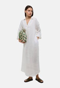 Robe en lin blanc avec un col et des manches trois-quarts, accompagnée d'un sac tissé vert et beige, et de sandales marron.
