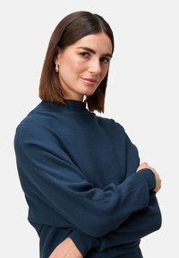 Granatowy sweter prążkowany z wysokim dekoltem i długimi rękawami, o luźnym kroju i miękkiej fakturze. Modelka pozuje z założonymi na siebie rękami.