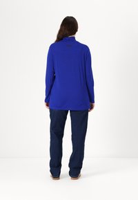 Langermet topp med høy hals i livlig blått stoff, kombinert med mørkeblå jeans. Har et enkelt design med en logo på baksiden.