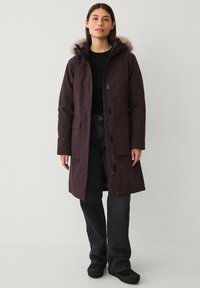 Donkerbruine parka met een voering van nepbont in de capuchon, een rits aan de voorkant en drukknoopsluitingen, voorzien van zijzakken en een lengte tot aan de knie.