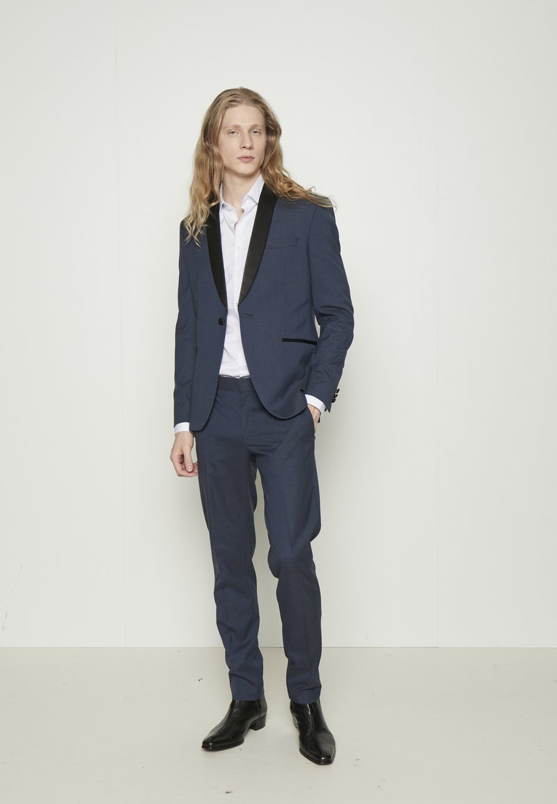 Completo blu navy con colletto a scialle nero, caratterizzato da una chiusura a un bottone, pantaloni slim-fit e stivaletti lucidi neri. Camicia bianca sotto.