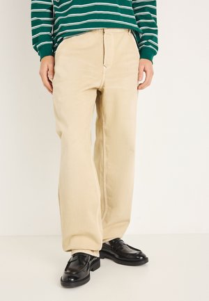 Pantaloni beige a gamba larga realizzati in cotone con una trama liscia e cuciture visibili. Abbinati a scarpe nere in pelle.