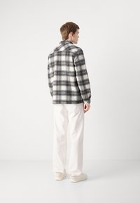 Plaid wool jacket in gray and white, with a collar and long sleeves. Worn over white pants and light sneakers, viewed from the back.