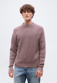 Νεαρός άνδρας με κατσαρά μαλλιά, ντυμένος με πουλόβερ από υφασμένο μάλλινο ύφασμα σε αποχρώσεις μοβ και ανοιχτό μπλε τζιν, στέκεται μπροστά σε απλό λευκό φόντο.