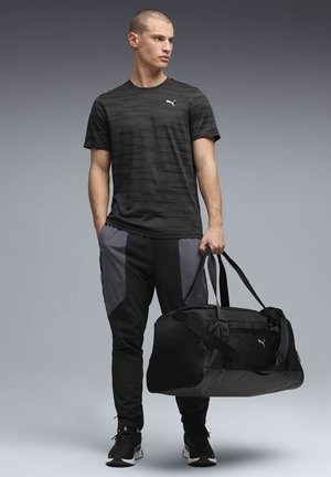 Atletisk man i svart Puma t-shirt och byxor håller en stor svart Puma-duffelväska, stående mot en grå gradientbakgrund.