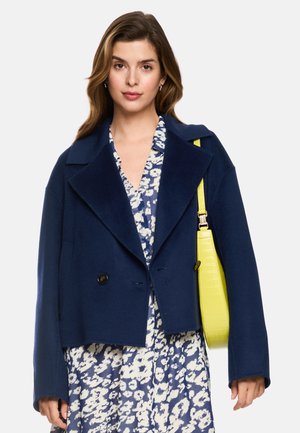 Frau trägt einen marineblauen Mantel über einem blau-weißen Blumenkleid und trägt eine leuchtend gelbe Schultertasche.