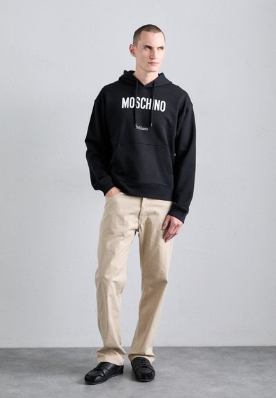 Hanorac negru cu textul "Moschino Milano", buzunar frontal, pantaloni bej și pantofi negri cu o suprafață texturată. Design simplu, croială lejeră.