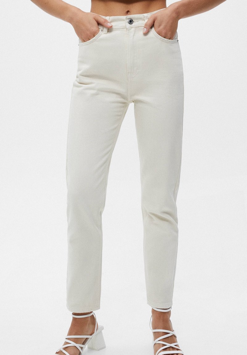 Jeans de mezclilla de algodón de color beige claro, de cintura alta, con un diseño de pierna recta, cinco bolsillos y cierre de botón. Estilizados con unos tacones blancos con tiras.