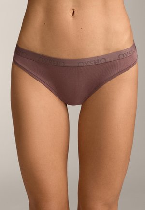 Frau trägt fliederfarbene Low-Rise-Hipster-Slips mit gebrandetem Bund auf beigem Hintergrund, zeigt Bauch und obere Oberschenkel.