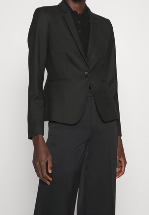 Persona con una blazer negra de un solo botón hecha a medida sobre una camisa negra con cuello y pantalones negros de pierna ancha contra un fondo liso.