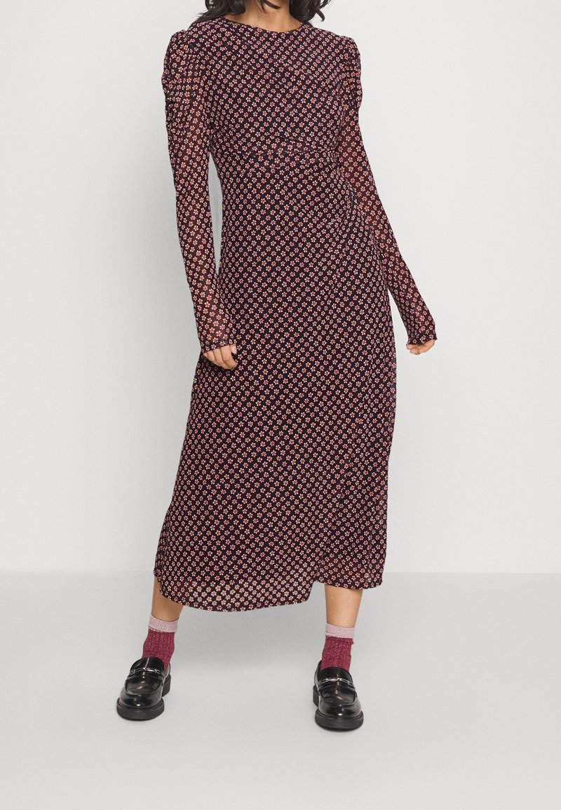 Femme portant une robe noire à manches longues et longueur cheville avec un petit imprimé floral rouge, associée à des chaussettes rouges et des mocassins noirs.