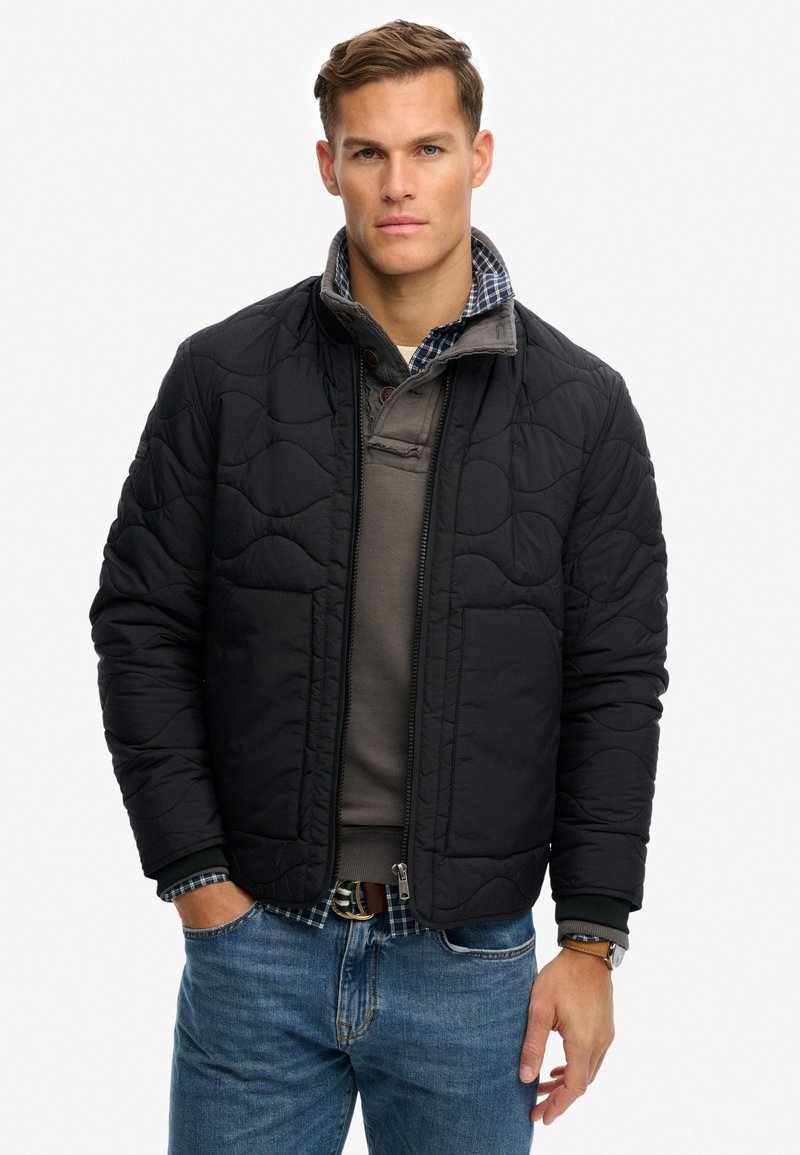 Czarna pikowana kurtka z zamkiem z przodu, ściągaczami na rękawach i kołnierzem, noszona na szarej koszulce. Do tego niebieskie dżinsy. Prosty, teksturowany design.