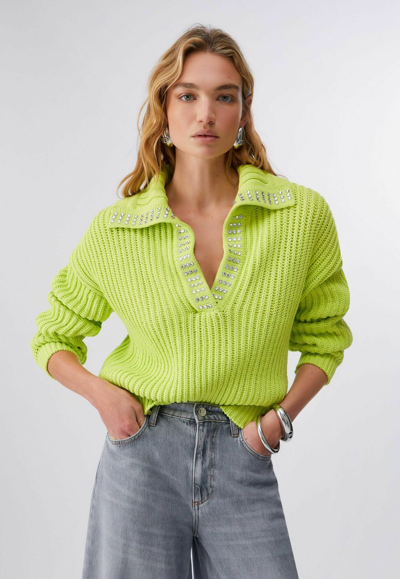 Femme aux longs cheveux blonds portant un pull en maille vert vif orné de strass, un jean taille haute gris, un bracelet argenté et des boucles d'oreilles.