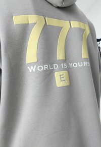 Grijze hoodie met grote gele cijfers "777", met de tekst "WORLD IS YOURS" in wit eronder en een gele accentvierkant met een logo.