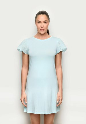 Frau mit zurückgebundenen Haaren, die ein hellblaues Kleid mit kurzen Ärmeln und Lochspitzen-Details an den Ärmeln trägt, steht vor einem einfarbigen Hintergrund.