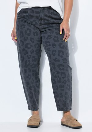 Person iført sorte bukser med leopardprint, beige slip-on sko, hvid top, gul neglelak og sølvringe, der står op ad en hvid væg.