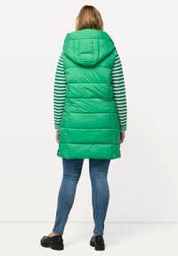 Grüne gepolsterte Weste mit Kapuze, die eine gesteppten Struktur und vertikale Nähte aufweist. Trägt man über einem gestreiften Langarmshirt und kombiniert mit blauen Jeans.