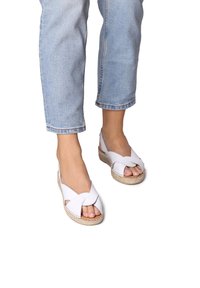 Sandalias blancas con tiras y un diseño cruzado, con una suela de esparto texturizada, punta abierta y un ligero tacón en cuña.