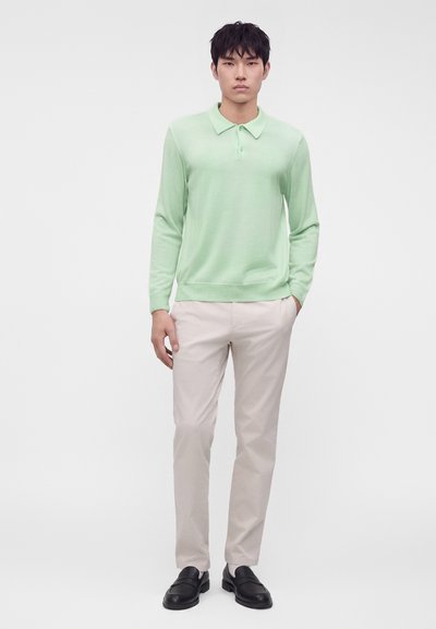 Jeune homme debout portant un polo vert clair à manches longues, un pantalon beige et des chaussures noires sur un fond blanc uni.