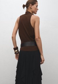 Haut marron sans manches avec un col montant, associé à une jupe noire à volants. Accessoires : larges bracelets marron et ceinture en cuir.