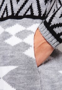 Grå stickad tröja med svart och vit geometrisk mönster, med en sidoficka och texturerade ärmslut. Mjuk, varm material.