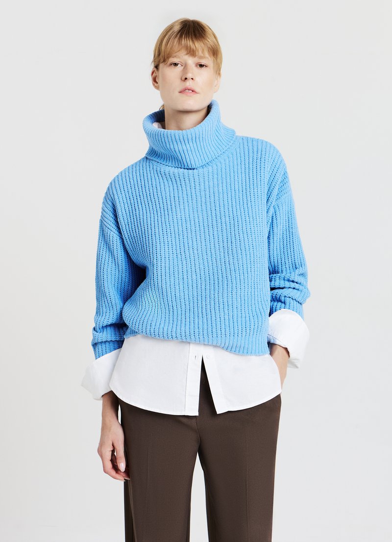 Hellblauer, gerippter Strickpullover mit hohem Kragen, über einem weißen, knöpfbaren Hemd getragen. Kombiniert mit braunen, weit geschnittenen Hosen.