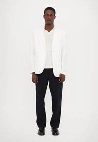 Weißer Leinenblazer über einem hellgrauen Pullover und navyfarbener Hose, ergänzt durch schwarze Schuhe. Das Design zeichnet sich durch klassische Revers und eine maßgeschneiderte Passform aus.