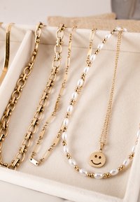 Cztery naszyjniki w kolorze złotym na beżowym ekspozytorze. Charakteryzują się grubymi ogniwami, perełkowymi zdobieniami oraz złotym wisiorkiem z uśmiechniętą buźką.
