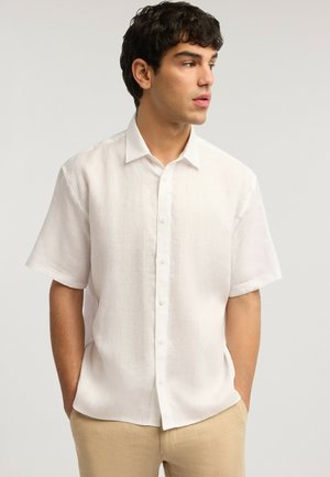 Junger Mann mit dunklem lockigem Haar, trägt ein weißes kurzärmeliges Hemd mit Knöpfen und beige Hosen, Hände in den Taschen, blickt zur Seite.